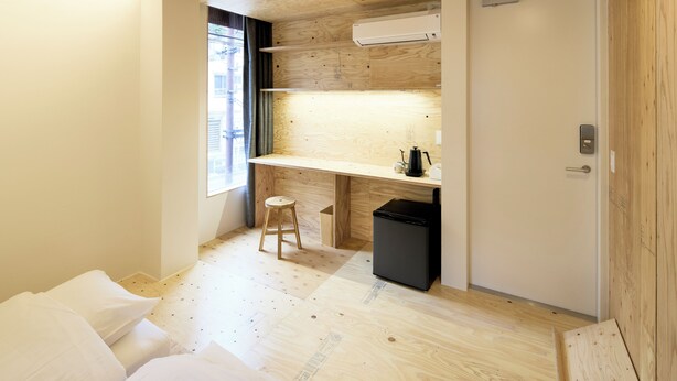 A compact room with a wooden floor, a small stool, a black trash bin, a white door, and a window with a view.