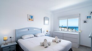 A neatly made bed with white linens and towels, a bedside table with a lamp, and a window with a view of a coastal town.