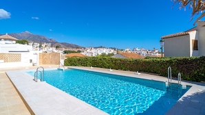 A rectangular swimming pool with clear blue water, surrounded by a tiled deck. In the background, there are buildings and a mountain range under a clear sky.