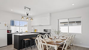 A modern kitchen with a central island, white cabinetry, and a dining area with a wooden table and white chairs. - C-Views ~ Aldinga Beach - Welcome To Your Dream Holiday House In Aldinga Beach (Aldinga Beach)