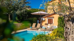 A pool area with lounge chairs and an umbrella, surrounded by greenery and a building with a tiled roof. - Holiday house  with pool,  garden, trampoline and a volleyball court (Vižinada)