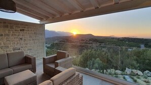 A balcony with wicker furniture and a stone wall, overlooking a mountainous landscape at sunset.