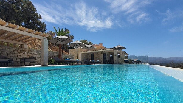 A clear blue pool with lounge chairs and umbrellas, surrounded by stone walls and a pergola.