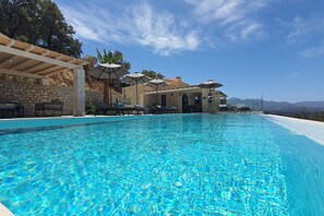 Une piscine d’un bleu éclatant, bordée de chaises longues et d’ombrelles, et entourée de murs en pierre et d’une pergola.