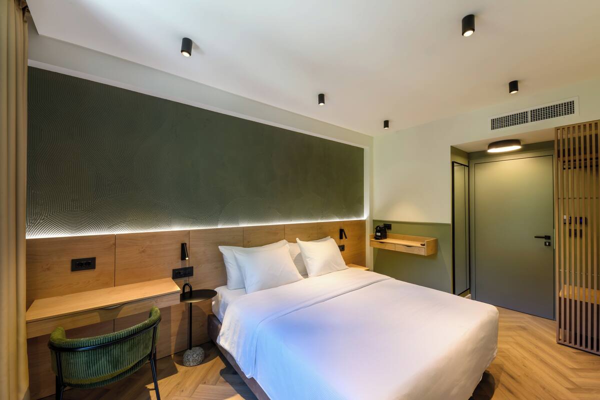 A modern hotel room with a large bed, a desk, and a chair. There is a wooden headboard, a wall-mounted shelf, and a door with a sliding panel.
