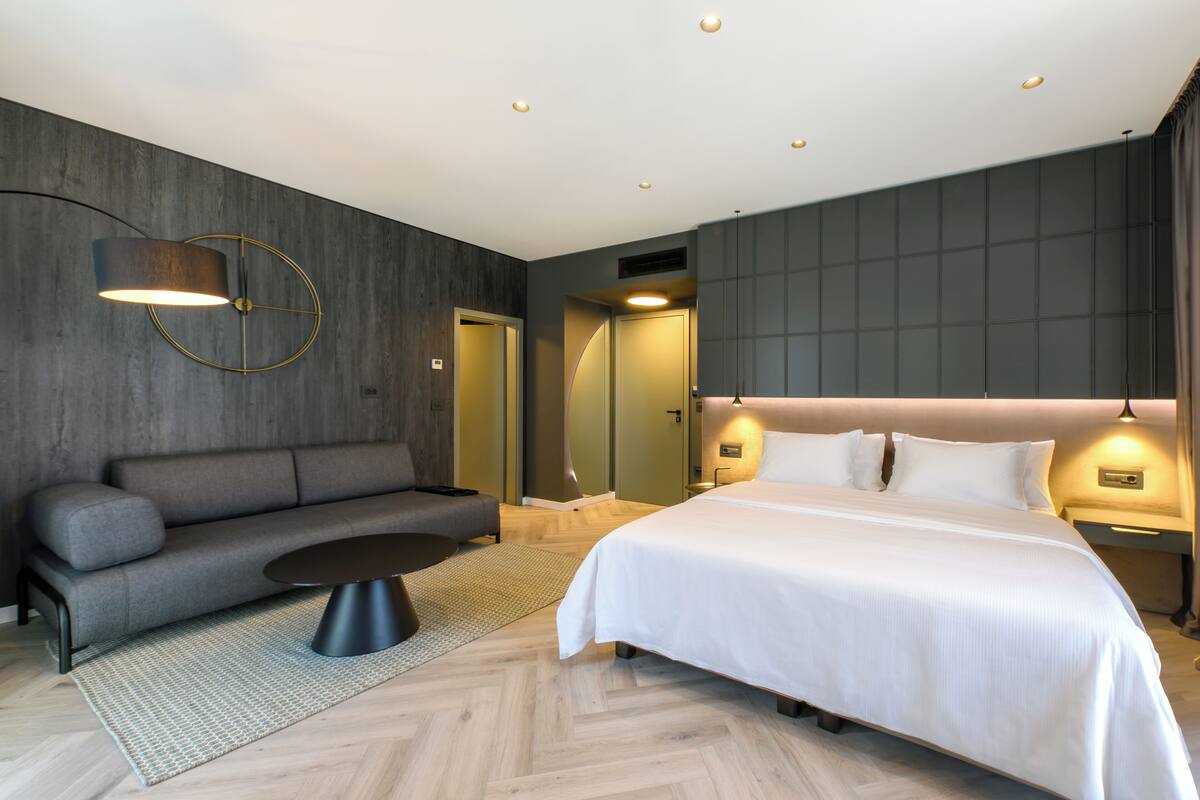 A modern hotel room with a large bed, a sofa, a coffee table, and a wall-mounted lamp.