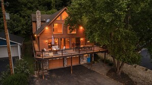 Eine zweistöckige Holzhütte mit einer Terrasse, einem Steinfundament und einer Garage.