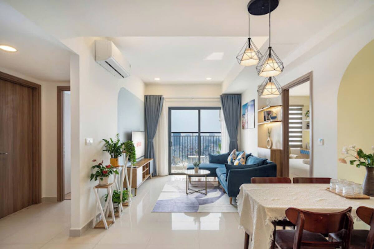 A modern living room with a sofa, dining table, and chairs. There is a balcony with a view, and the room has a large window.