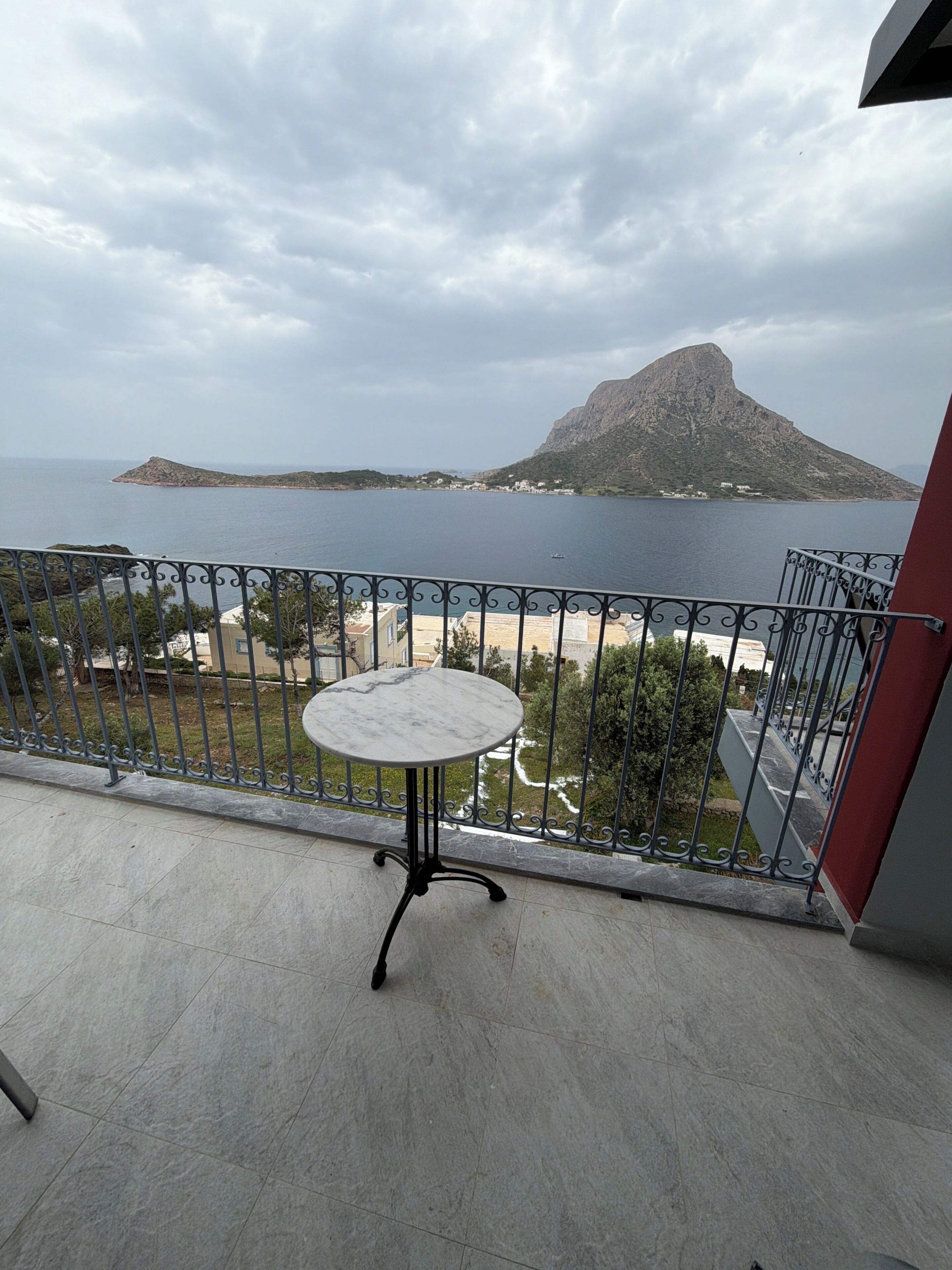 Ein Balkon mit einem Marmortisch, einem Metallgeländer und Blick auf eine Küstenlandschaft mit einem Berg.