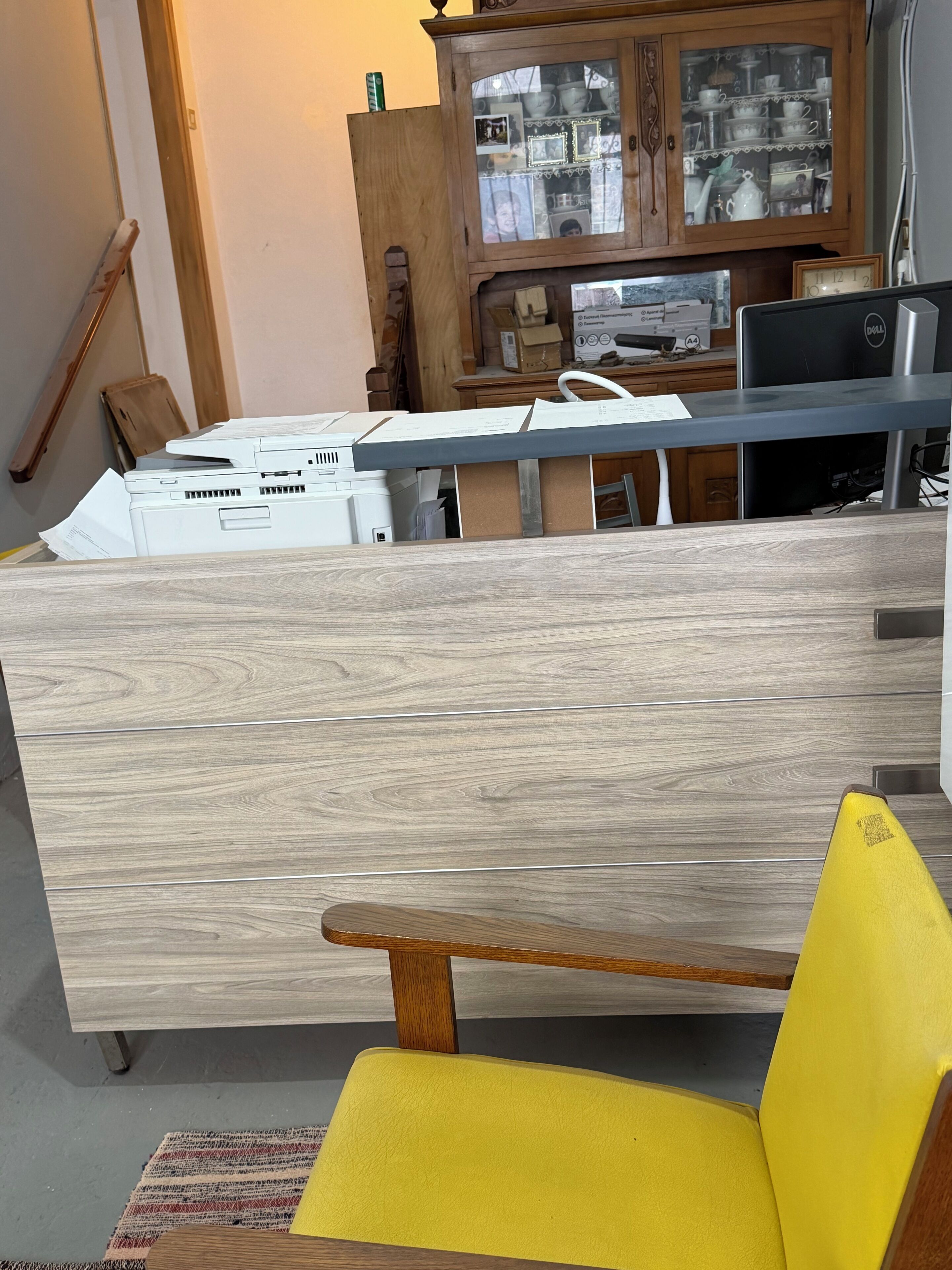 Een kamer met een houten kast vol diverse spullen, een bureau met papieren, een gele stoel en een Dell computer.