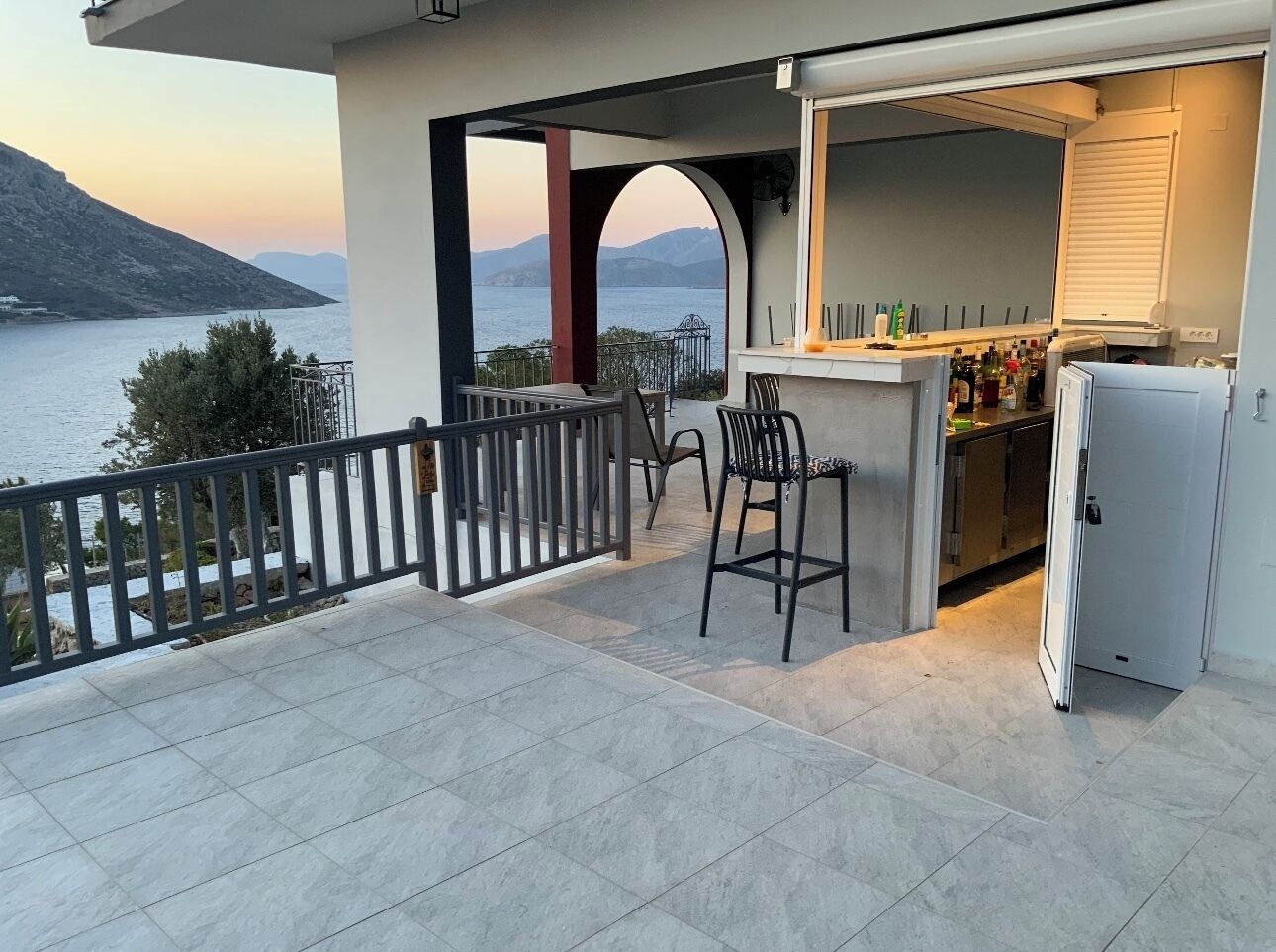 Un balcón con vista al mar, una zona de bar con sillas y un refrigerador.