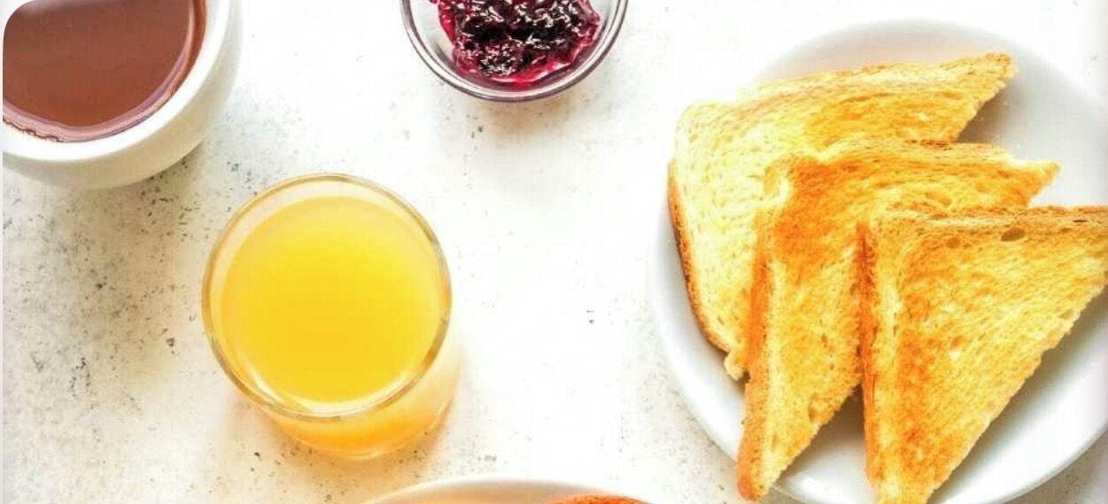 Desayuno con tostadas, mermelada y jugo de naranja sobre una mesa blanca.