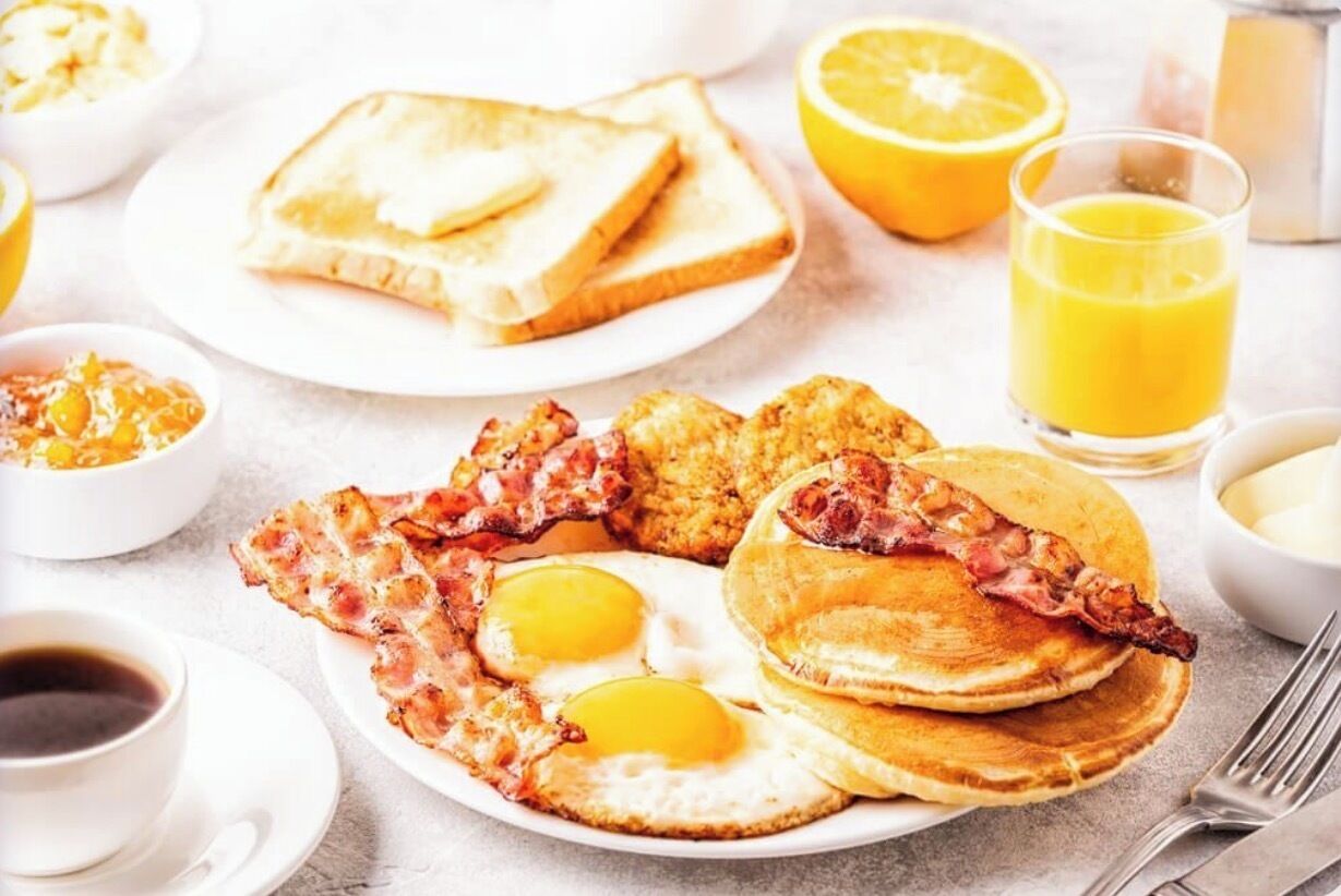 Prato de café da manhã com ovos, bacon e panquecas, acompanhado de torradas, suco de laranja e café.