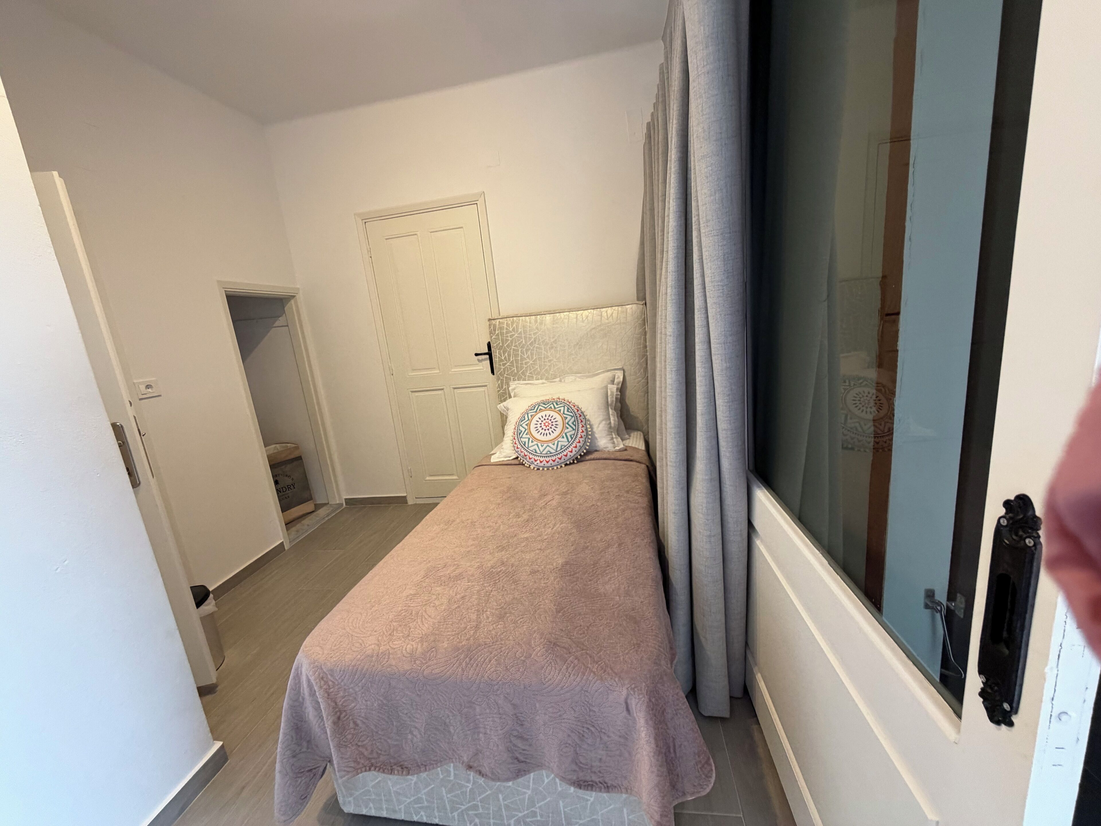 A bedroom with a bed, a door, a window with curtains, and a wall-mounted light fixture.