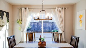 A dining room with a wooden table, chairs, a chandelier, and a view of a wooden deck through a window with curtains. - Charming Retreat: King Bed, Full Kitchen & Spacious Backyard. Perfect Location! (Hillsboro)