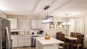 A modern kitchen with white cabinets, a stainless steel refrigerator, a microwave, and a dishwasher. There is a dining area with a wooden table and chairs. - Charming Retreat: King Bed, Full Kitchen & Spacious Backyard. Perfect Location! (Hillsboro)