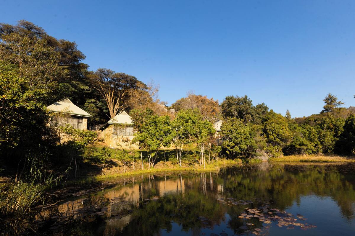 A serene lakeside setting with a traditional house and a small building, surrounded by dense trees and a calm water body reflecting the structures.