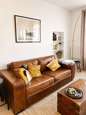 A living room with a brown leather sofa, a framed picture on the wall, a small shelf with decorative items, and a wooden side table with a board game.