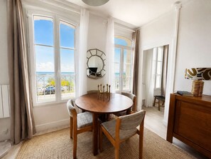 Ein Essbereich mit rundem Tisch und Stühlen, großen Fenstern mit Vorhängen sowie Blick aufs Meer und einer Holzkabine.