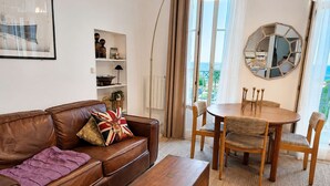 A living room with a brown leather sofa, a wooden coffee table, and a dining table with chairs. There is a bookshelf, a floor lamp, and a large mirror on the wall.