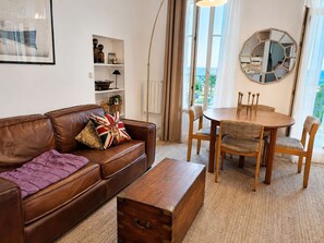 A living room with a brown leather sofa, a wooden coffee table, and a dining table with chairs. There is a bookshelf, a floor lamp, and a large mirror on the wall.
