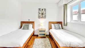 Un dormitorio con dos camas individuales, una mesita de noche con lámpara, un cuadro en la pared y una ventana con cortinas.