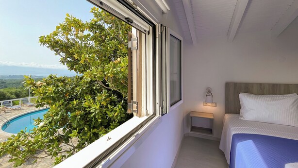 Un dormitorio con un ventanal grande, una cama, una mesita de noche con lámpara y vistas a una piscina y follaje.
