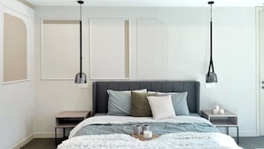 Un dormitorio moderno con una cama tapizada en gris, ropa de cama blanca y gris, dos mesitas de noche con lámparas y un amplio ventanal con persianas.