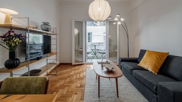 Un salon moderne avec un canapé, une table basse, un téléviseur posé sur une étagère et une grande fenêtre donnant sur l’extérieur.