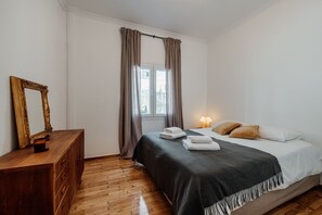 Ein Schlafzimmer mit einem hölzernen Schminktisch, einem Bett mit dunkler Tagesdecke und zwei Kissen, einem Fenster mit Vorhängen und einer Lampe auf dem Nachttisch.