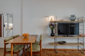 Ein modernes Wohnzimmer mit einem Holzesstisch, grün gepolsterten Stühlen, einem Fernseher auf einem Metallständer, einer Blumenvase und einer Schale mit Orangen auf dem Tisch.