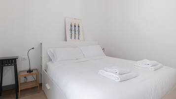 Una cama bien hecha con sábanas y toallas blancas, una mesita de noche de madera con una lámpara y un cuadro en la pared.