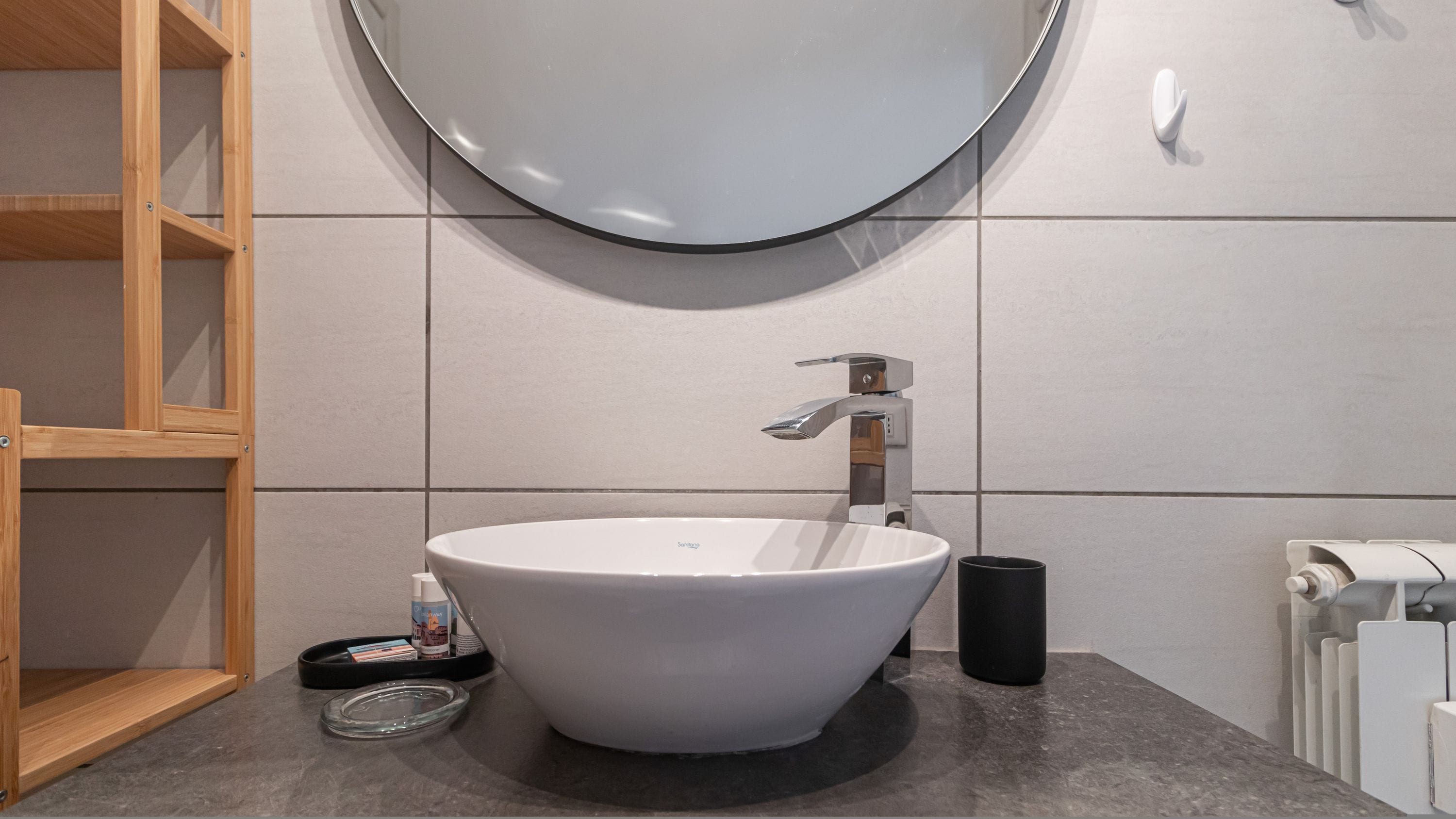 A modern bathroom with a large white sink, a round mirror, and a wooden shelf.