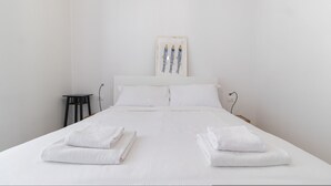 Un lit bien fait, avec du linge de lit et des serviettes blanches, un petit tabouret noir et une œuvre d’art encadrée au mur.