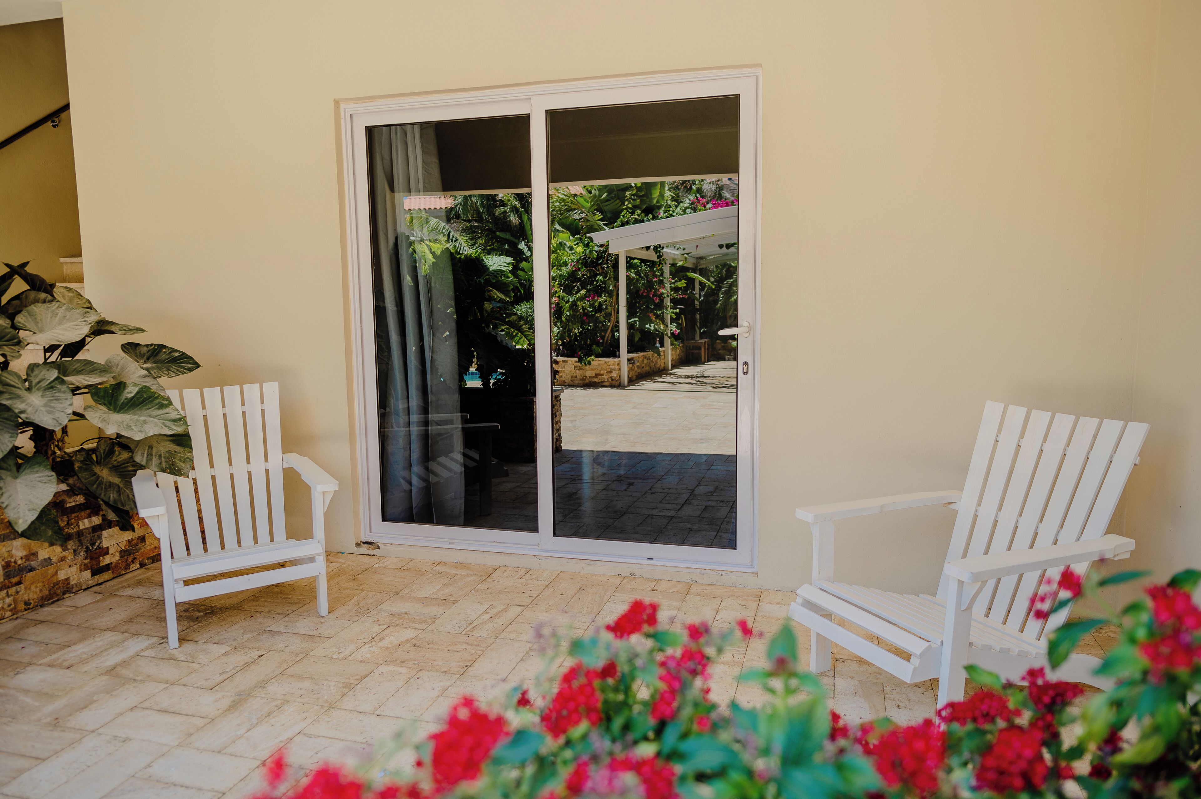 A patio area with two white Adirondack chairs, a large sliding glass door leading to a covered outdoor space, and a potted plant with large leaves.