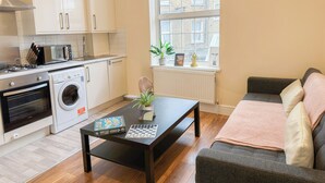A compact kitchen with a stove, microwave, and washing machine, a window with a view of buildings, a black table with a board game and a plant, and a grey sofa with a blanket. - 2BDR flat in Hammersmith with Wi-Fi & Workspace near Shepherd's Bush & Westfield (London)