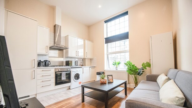 A modern kitchen with white cabinets, a stainless steel range hood, and a built-in oven. There is a gray sofa and a small table with a plant and a sign in the living area. - 2BDR flat in Hammersmith with Wi-Fi & Workspace near Shepherd's Bush & Westfield (London)