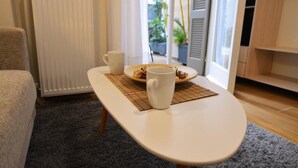 A small, round table with two white cups and a plate of food, positioned in a room with a radiator, a window with blinds, and a grey carpet.