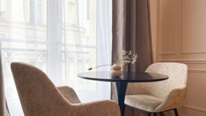 Une pièce avec deux fauteuils beiges disposés autour d’une petite table ronde, un vase rempli de fleurs et une vue sur un bâtiment à travers des rideaux transparents.
