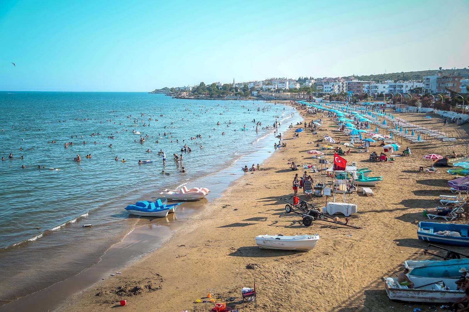 Een strand met veel mensen die zwemmen en zonnebaden, boten en strandparasols.