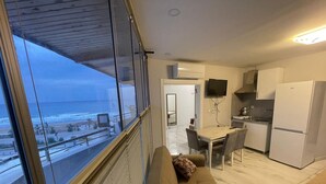 A modern living room with a large window overlooking the sea, a sofa, and a dining area with a kitchen in the background. - BeyazmotelRestaurant (Erdemli)
