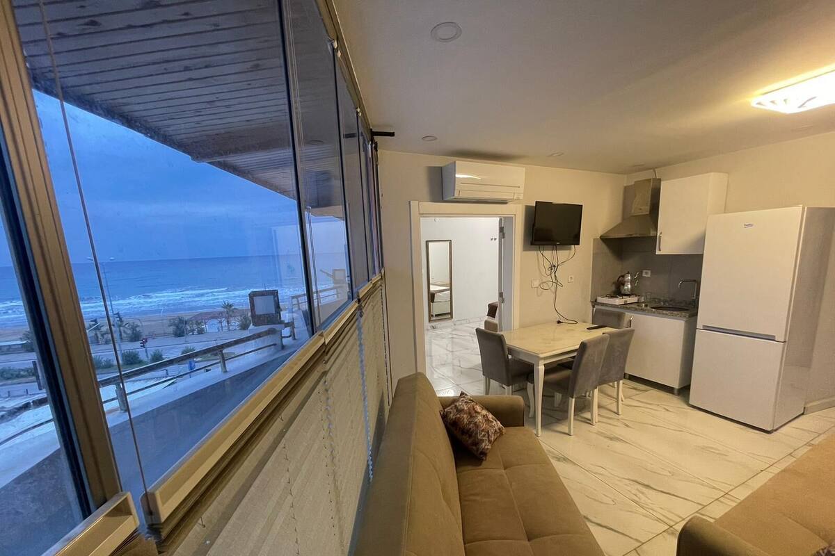 Una sala de estar moderna con un ventanal grande que da al mar, un sofá y un comedor con una cocina al fondo.
