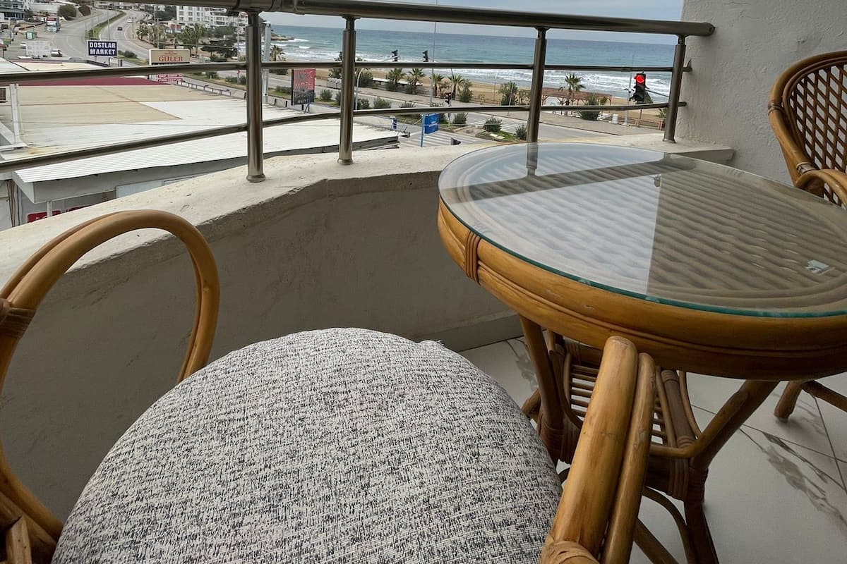 Un balcón con una mesa de vidrio y sillas de mimbre, con vista a una playa y al paisaje urbano.
