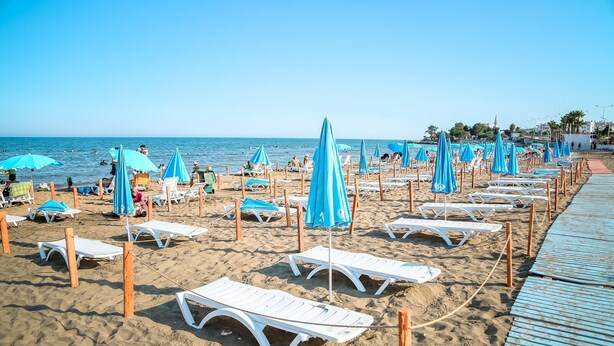 A beach with rows of white sun loungers and blue umbrellas, a wooden walkway, and a clear view of the sea. - BeyazmotelRestaurant (Erdemli)