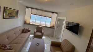A modern living room with a sofa, armchair, and a coffee table. There is a large window with blinds, a flat-screen TV mounted on the wall, and a ceiling light fixture. - BeyazmotelRestaurant (Erdemli)