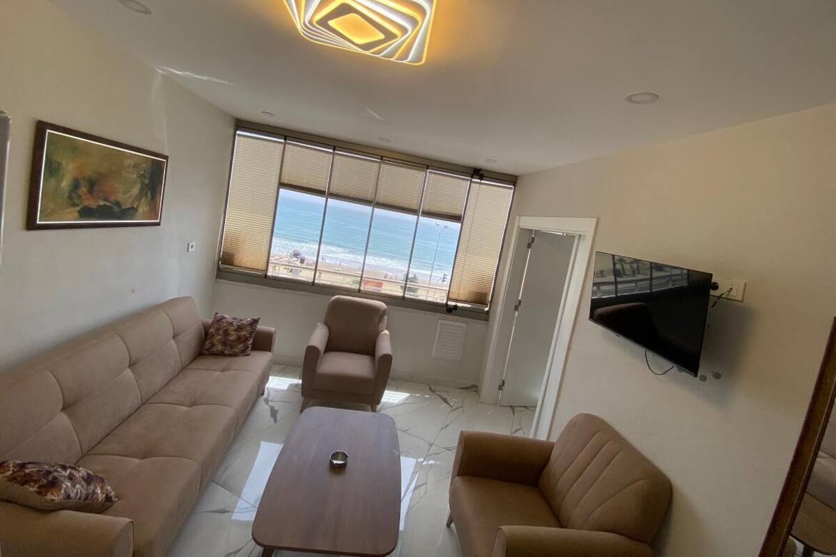 Una sala de estar moderna con sofá, sillón y una mesa de centro. Se ve un ventanal con persianas, un televisor de pantalla plana montado en la pared y una lámpara en el techo.