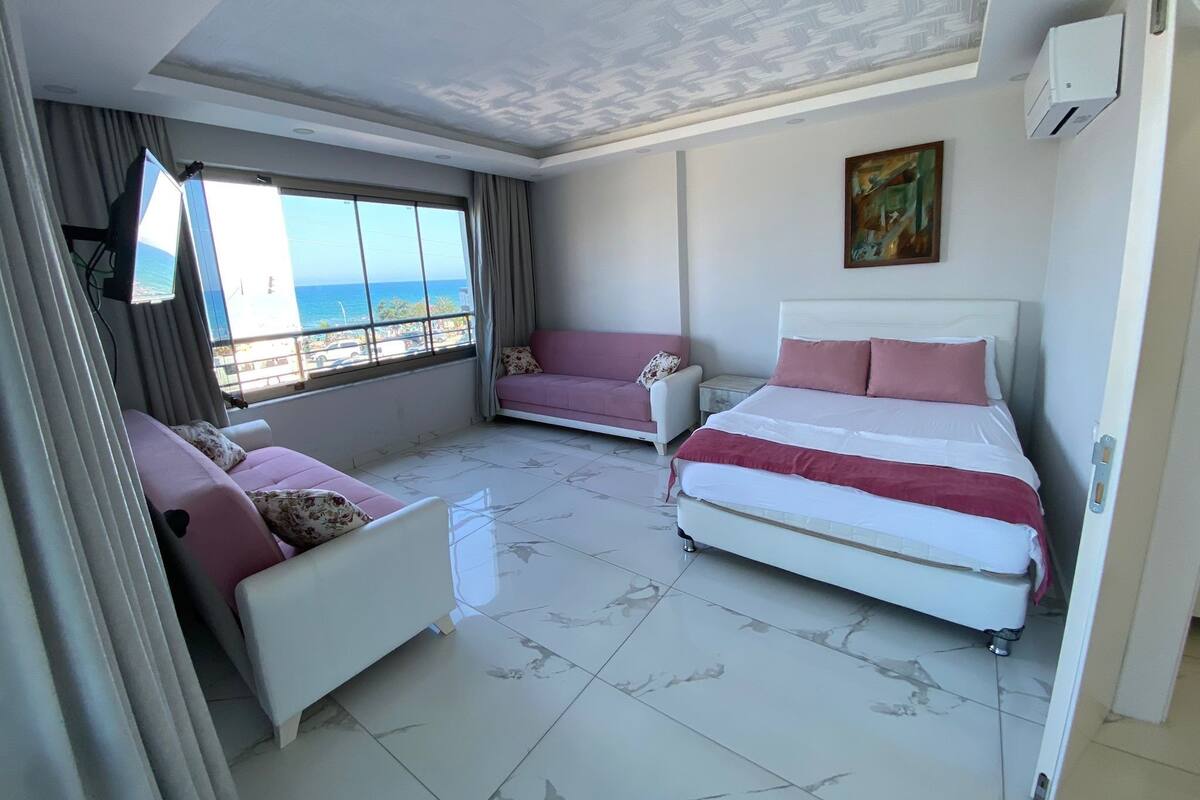 Un dormitorio con cama, sofá y una mesita. Se ve una ventana con vista al mar.