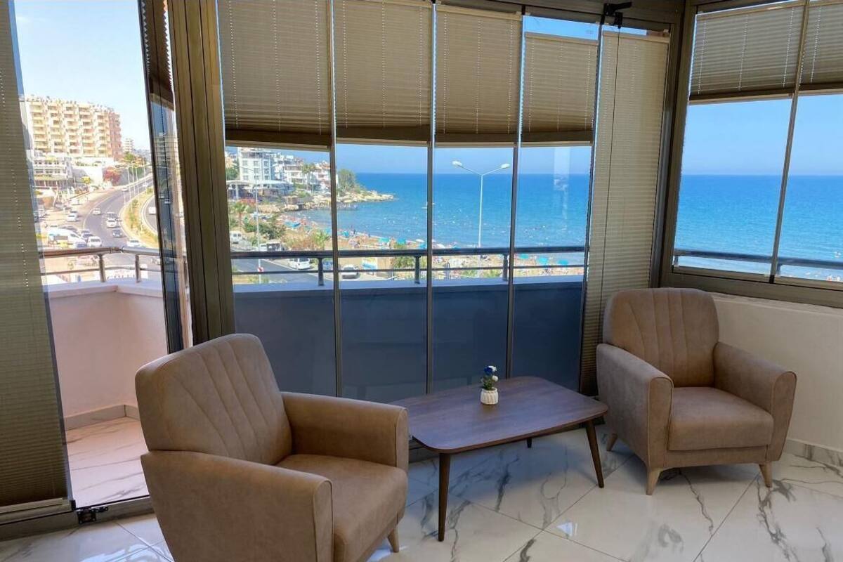 Un balcón con dos sillones beige y una pequeña mesa de madera, con vista a un paisaje urbano costero que incluye edificios y el mar.