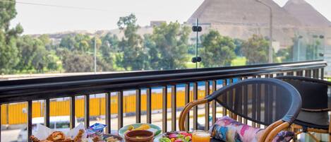A balcony with a table set for breakfast, including croissants, eggs, and a glass of orange juice, overlooking a pyramid.