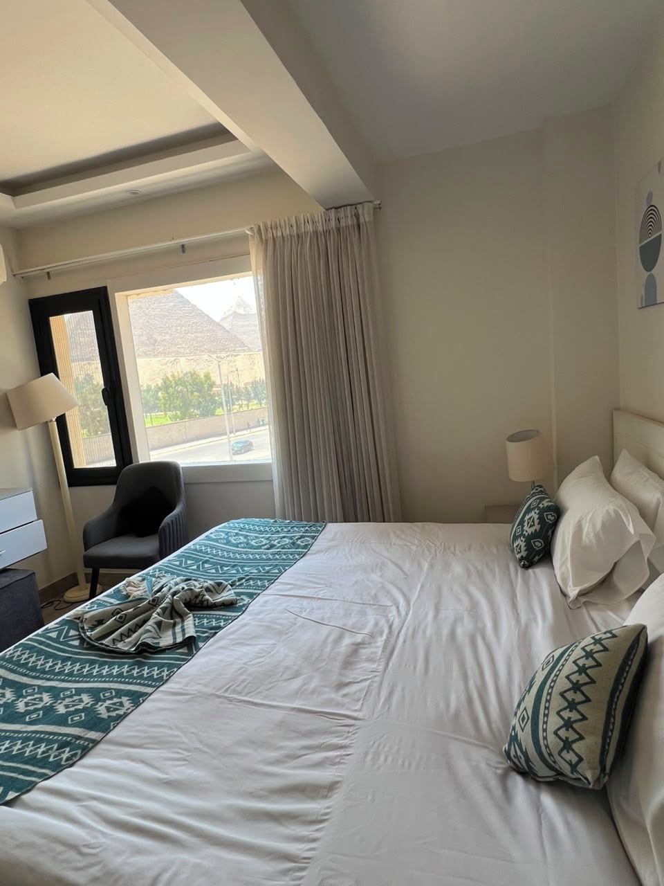 Habitación de hotel con cama, una silla, escritorio, lámpara y vistas al exterior.